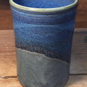 May include: A handmade ceramic tumbler with a blue and gray glaze. The top rim has a light green accent. The tumbler has a cylindrical shape and is suitable for beverages. The glaze has a textured appearance.