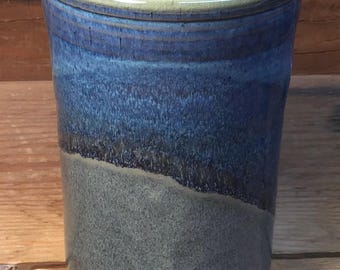 May include: A handmade ceramic tumbler with a blue and gray glaze. The top rim has a light green accent. The tumbler has a cylindrical shape and is suitable for beverages. The glaze has a textured appearance.