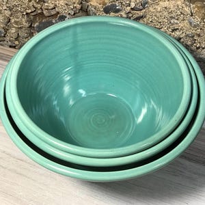 May include: A set of two nested ceramic bowls with a smooth, glossy finish in a vibrant turquoise color. The bowls feature a rounded shape with a consistent, solid color glaze throughout the interior and exterior surfaces, showcasing a simple design.