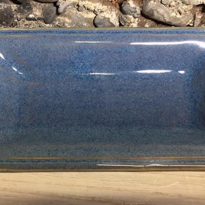May include: A rectangular ceramic tray with a glossy blue glaze and a speckled finish. The tray features slightly raised edges and a smooth, reflective surface with subtle tan-colored trim along the outer border of the rim.