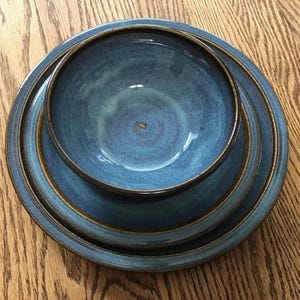 May include: A set of three stacked ceramic dishes consisting of a large dinner plate, a smaller salad plate, and a deep bowl. Each piece features a mottled blue glaze with dark brown edges and a speckled texture throughout the surface finish.