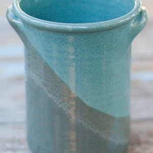 May include: A handcrafted ceramic utensil holder in shades of teal and gray. The cylindrical holder features a diagonal color block design and small handles on either side. The interior is a vibrant teal, and the exterior has a textured finish.
