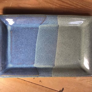 May include: A rectangular ceramic tray featuring a smooth, glazed finish with a color gradient transitioning from a deep periwinkle blue on the left side to a muted, earthy sage green on the right side, separated by a textured central band.