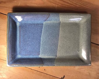 May include: A rectangular ceramic tray featuring a smooth, glazed finish with a color gradient transitioning from a deep periwinkle blue on the left side to a muted, earthy sage green on the right side, separated by a textured central band.