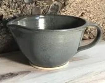Può includere: Una ciotola per mescolare in ceramica grigio scuro con finitura lucida, caratterizzata da un corpo ampio e arrotondato, un manico laterale singolo e un piccolo beccuccio sul bordo. La base della ciotola non è smaltata, rivelando un bordo inferiore circolare beige chiaro.