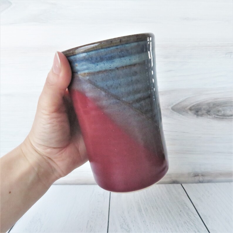 May include: A handmade ceramic mug with a red and blue glaze. The mug has a wide, rounded base and a slightly flared lip.