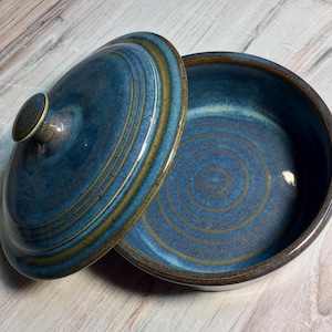 May include: A round ceramic lidded bowl featuring a deep blue glaze with concentric circular patterns in a muted golden-brown hue. The bowl has a matching lid with a small central knob, and both pieces exhibit a smooth, glossy finish throughout.