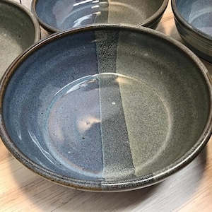 May include: A set of round ceramic bowls featuring a two-tone glaze design. Each bowl displays a distinct vertical split with one side in a deep blue hue and the other in a muted grey-green tone, finished with a smooth, glossy surface texture.