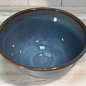 May include: A handmade ceramic bowl with a deep blue glaze and a contrasting brown rim. The bowl has a smooth, glossy finish and a wide, open design. This kitchenware item is perfect for serving salads, soups, or other dishes.