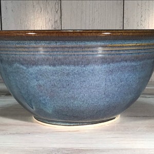 May include: A large, handmade ceramic bowl with a wide, rounded shape. The bowl features a gradient of blue hues, transitioning from a darker shade at the top to a lighter tone towards the base. The rim is a rich brown, and the base is white.
