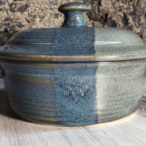 May include: A ceramic casserole dish with a lid. The dish is divided into two vertical sections, one in a deep blue and the other in a muted gray-green. The lid has a small, rounded handle. The dish is suitable for baking and serving.