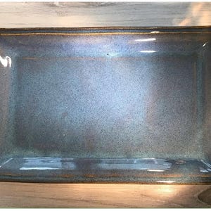 May include: Rectangular ceramic serving dish with a glossy, speckled blue-gray surface and a dark brown rim. The dish has slightly rounded corners and a smooth, reflective finish, suitable for serving food.