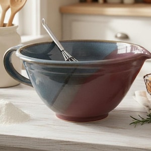 May include: A ceramic mixing bowl featuring a two-tone design with a deep blue upper section and a dark red lower section. The bowl includes a side handle and a pouring spout, with a stainless steel wire whisk resting inside the basin.