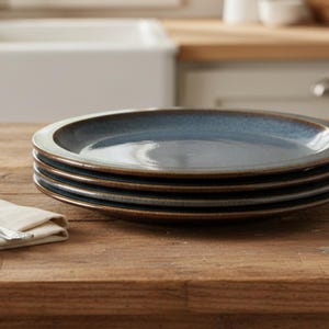 May include: A stack of four round ceramic dinner plates with a glossy blue center and a dark brown rim. The plates have a smooth, reflective surface and are arranged in a neat pile. A silver fork rests on a folded white cloth napkin nearby.