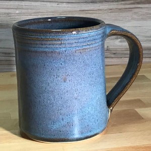 May include: A handmade ceramic mug with a blue glaze and a brown rim and handle. The mug has a cylindrical shape and a large handle for easy gripping. The mug is suitable for hot or cold beverages.