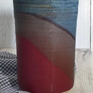 May include: A ceramic utensil holder with a blue, gray, and red glaze. The holder is cylindrical and has a wide opening at the top.