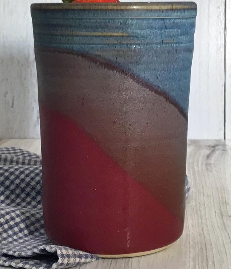 May include: A ceramic utensil holder with a blue, gray, and red glaze. The holder is cylindrical and has a wide opening at the top.