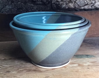 May include: A set of two nested ceramic bowls featuring a tri-color glaze design. The exterior and interior surfaces are divided into diagonal bands of bright turquoise, muted sage green, and dark charcoal gray, with a white base rim.