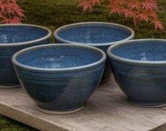 May include: A set of four round ceramic bowls with a deep blue speckled glaze. Each bowl features a smooth, curved interior and a series of subtle horizontal ridges encircling the exterior wall, finished with a light-colored rim and base.