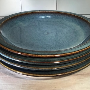 May include: A stack of four round ceramic dinner plates featuring a speckled blue-grey center and a dark brown rim. The plates have a smooth, glazed finish with a slightly raised edge, showcasing a consistent color pattern across the set.