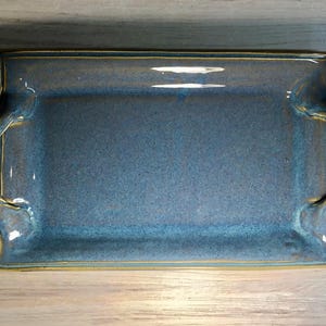 May include: A rectangular ceramic serving tray with a glossy blue glaze and a speckled finish. The tray features two integrated, twisted handles on the left and right sides, with visible yellow-toned edges along the rim and handle details.