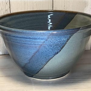 May include: A large, round ceramic bowl featuring a glossy glaze with a distinct diagonal color split. One side of the bowl is a deep, rich navy blue, while the other side transitions into a muted, earthy sage green. The rim is dark and smooth.