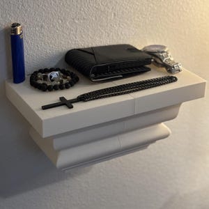 May include: A collection of accessories displayed on a white shelf. Items include a blue lighter, a black textured wallet, a silver wristwatch, a beaded bracelet, a black cross pendant, and a chain necklace.