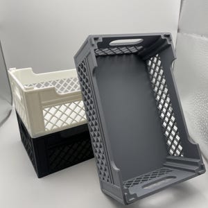 May include: Three plastic crates, one gray, one black, and one white, stacked on top of each other. The crates are made of a grid-like material and have a handle on the top.