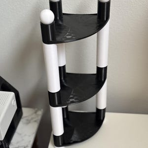 May include: A three-tiered corner shelf with a modern design. The shelves are black, and the supports are white with black accents. Each support is topped with a white sphere. The shelf is made of plastic and is designed to fit in a corner.