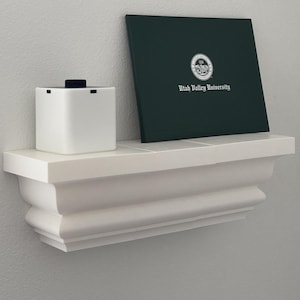 May include: A white, wall-mounted shelf with a decorative, curved design. On the shelf are a white square object with a black top, and a dark green diploma case with the text "Utah Valley University" and a university seal.