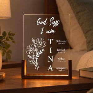 May include: A rectangular clear acrylic plaque with a warm light base, featuring white floral line art and the text God Says I am TINA, with the words Delivered, Invited, Noble, and Accepted alongside corresponding Bible verse references.