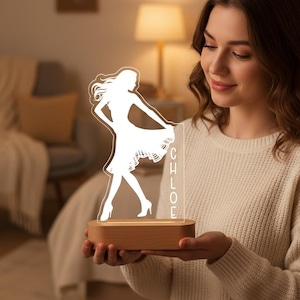 Op de afbeelding: LED-lamp met een houten voet, met een witte silhouet van een vrouw in een jurk. De naam "CHLOE" staat verticaal afgedrukt. De lamp straalt een warm licht uit en wordt door een persoon vastgehouden.