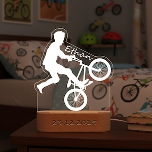 Op de afbeelding: Een gepersonaliseerd nachtlampje met een wit silhouet van een persoon op een BMX-fiets. De naam "Ethan" staat op de fiets en de datum "23.12.2025" is gegraveerd op de houten basis. De lamp staat op een donkere houten tafel.