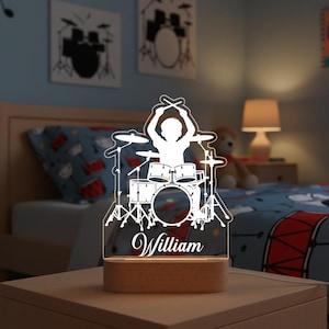 May include: A white acrylic drum set night light with the name "William" engraved on the base. The lamp features a silhouette of a drummer playing a drum set. The base is made of wood. The background includes drum-themed artwork.