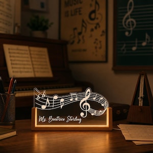 May include: A rectangular wooden base supports a clear acrylic panel illuminated with a warm light. The panel features a white etched design of a musical staff with notes and a treble clef, with the text Ms. Beatrice Sterling printed below.