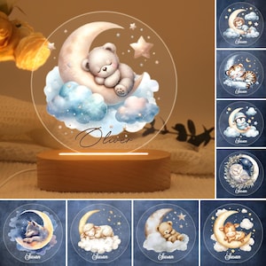 Może przedstawiać: Lampka nocna z przezroczystą podstawą akrylową i drewnianą podstawą. Panel akrylowy przedstawia sierp księżyca ze śpiącym zwierzęciem na nim. Na panelu napisano imię "Oliver". Lampka nocna jest otoczona innymi lampkami nocnymi z różnymi zwierzętami na nich.