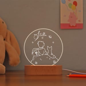 Pode incluir: Uma luz noturna com um design circular iluminado que retrata um personagem e uma raposa. O nome "Stella" está escrito em itálico. A luz é montada em uma base de madeira retangular.