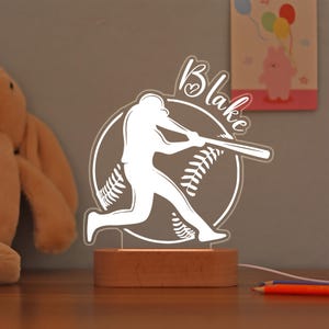 May include: A white acrylic baseball-themed night light with the name "Blake" in cursive. The design features a baseball player swinging a bat, set against a baseball. The light sits on a wooden base.