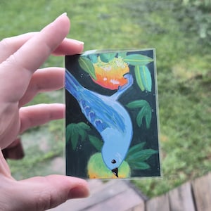 May include: A small, rectangular painting of a blue bird perched on a green and yellow fruit. The bird is painted with a light blue hue, and the fruit is a mix of yellow and green. The background is dark green with green leaves.