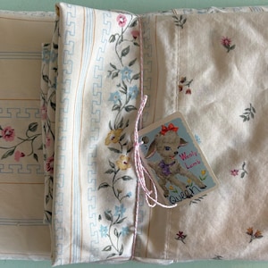 May include: Folded vintage queen-sized bed sheets with floral and striped patterns in cream, pink, blue, and yellow. A tag with a cartoon lamb is attached with pink string. The sheets are folded and ready for use.