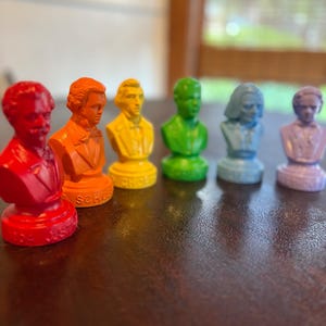 May include: A row of six colorful busts of historical figures. The busts are painted in a variety of colors, including red, orange, yellow, green, blue, and purple. The busts are arranged in a line on a brown surface.
