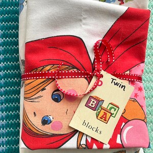 May include: A set of two white twin-sized bed sheets with a red and white print of a girl in a red hooded cloak. The sheets are tied together with a red ribbon and a tag that says "Twin" and "blocks" with colorful letters.