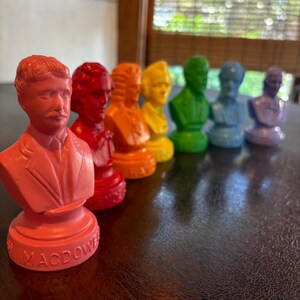 May include: A row of colorful busts, each depicting a historical figure. The busts are arranged in a gradient of colors, from red to purple. The base of the first bust reads "MACDOWE". The busts are on a dark brown surface.