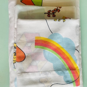 May include: A stack of three white cotton pillowcases with a colorful rainbow, cloud, and whimsical design. The design features a rainbow with red, orange, yellow, green, and blue stripes.