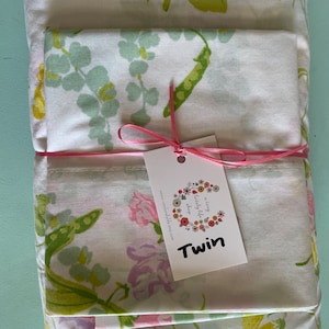 May include: A set of white twin-sized bed sheets with a floral pattern featuring green leaves, pink and yellow flowers, and pea pods. The sheets are folded and tied with a pink ribbon. A white tag with the text "a very lovely little shop" and "Twin" is attached to the ribbon.