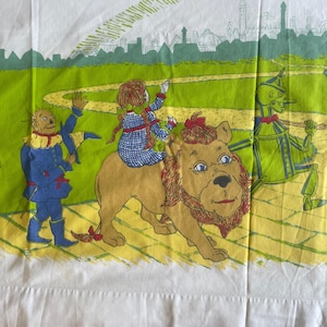 May include: A vintage cotton sheet featuring a scene from the Wizard of Oz. Dorothy, the Scarecrow, the Tin Man, and the Cowardly Lion are walking down the yellow brick road towards the Emerald City. The sheet is white with a green and yellow design.