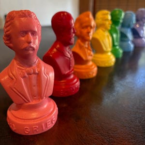 May include: A row of colorful busts of historical figures, each painted in a different color of the rainbow. The busts are arranged on a dark brown surface, with the first bust being a coral color. The word "Grieg" is visible on the base of the first bust.