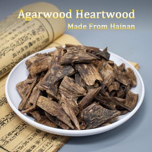 May include: A white bowl filled with dark brown, jagged pieces of Agarwood Heartwood. The text Agarwood Heartwood Made From Hainan is visible above the bowl, which sits on a piece of aged, light brown paper with black Chinese characters.
