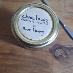 May include: A jar with a gold lid containing raw honey with clove buds essence extract. The label is handwritten. A silver metal tool is in the background, resting on a wooden surface.