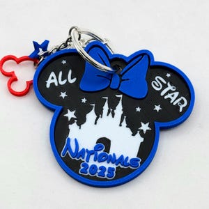 Könnte beinhalten: Blau-schwarzer und weißer Schlüsselanhänger in Form eines Mäusekopfes. Der Schlüsselanhänger trägt die Aufschrift "ALL STAR Nationals 2025" und eine Schlossgrafik. Ein roter Mickey Mouse-Kopf und ein blauer Stern-Anhänger sind befestigt.
